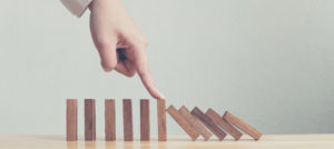 A hand tipping over a row of wooden dominoes, symbolizing the chain reaction of corporate governance decisions and risk management processes